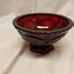 May include: A ruby red glass bowl with a pedestal base. The bowl has a scalloped rim and a diamond pattern on the sides. The bowl is likely vintage and suitable for serving fruit or candy.