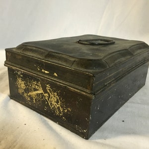 Könnte beinhalten: Eine antike schwarze Metallbox mit einem Scharnierdeckel und einem Metallgriff. Die Box hat einen abgenutzten, verwitterten Look, wobei etwas goldene Farbe durchscheint.