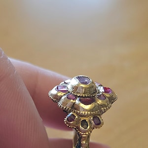 May include: A gold-toned ring with a floral design, featuring multiple red gemstones. The ring's setting has intricate detailing, and the gemstones are set in a cluster pattern. The ring is held between fingers.