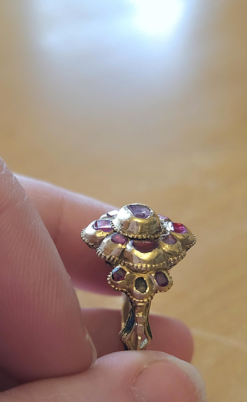 May include: A gold-toned ring with a floral design, featuring multiple red gemstones. The ring's setting has intricate detailing, and the gemstones are set in a cluster pattern. The ring is held between fingers.
