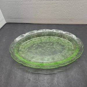 May include: An oval-shaped light green glass platter featuring a pressed floral and leaf pattern across the center and a scalloped edge. The translucent glass has a smooth surface with raised details that define the botanical design elements.