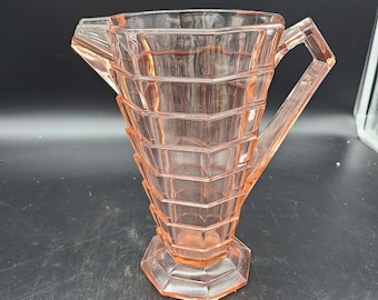 May include: A pink glass jug with a geometric design. The jug has a wide base and a narrow spout. The handle is attached to the side of the jug.