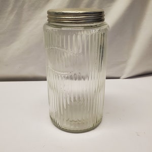 Op de afbeelding: Een heldere glazen pot met verticale geribbelde details en een zilveren metalen deksel. De pot heeft een cilindrische vorm en is leeg. De pot wordt waarschijnlijk gebruikt voor het bewaren van droge goederen.