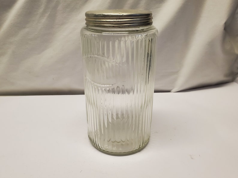 May include: A clear glass jar with vertical ribbed detailing and a silver metal lid. The jar has a cylindrical shape and is empty. The jar is likely used for storage of dry goods.