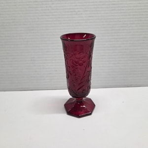 May include: A ruby red glass vase with a floral pattern. The vase has a flared top and a hexagonal base. The embossed floral design covers the entire body of the vase. The vase is approximately 7 inches tall.
