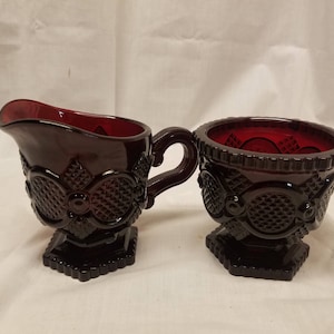 Peut inclure: Un ensemble de crémier et sucrier en verre rouge rubis. Le crémier a une anse et un bec verseur, tandis que le sucrier est ouvert. Les deux pièces présentent un motif texturé détaillé et reposent sur une base carrée. L'ensemble est probablement vintage.