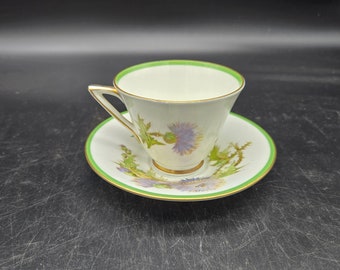 Peut inclure: Une tasse à thé et une soucoupe blanches avec un bord vert et un motif floral représentant des chardons violets et verts.
