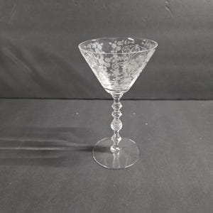 May include: A clear, etched glass cocktail glass with a floral design. The glass has a conical bowl, a decorative stem with a ball design, and a round base. The glass is likely used for serving cocktails or other beverages.