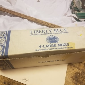 May include: A rectangular box labeled "LIBERTY BLUE" with "A Collection of Historic Colonial Scenes" printed on it. The box also says "4-LARGE MUGS" and "Staffordshire Ironstone Made in England". The box is light blue and white.
