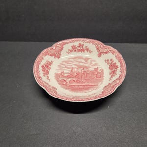 May include: A white and red ceramic bowl with a scalloped edge. The bowl features a red illustration of a castle scene in the center and floral patterns around the rim. The bowl is on a dark surface.