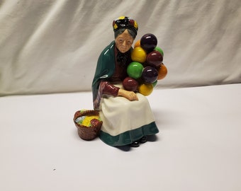 May include: A ceramic figurine of an elderly woman seated, holding a cluster of colorful orbs. She wears a green shawl, a white dress with a green trim, and a patterned head covering. A basket with colorful items sits beside her.