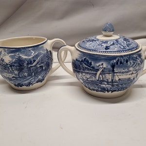May include: A cream-coloured creamer and lidded sugar bowl set with blue transferware scenes. The creamer has a spout and handle, while the sugar bowl features two handles and a knobbed lid. The blue and white design depicts landscapes and a steamboat.
