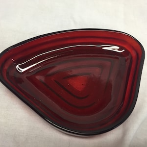 May include: A triangular-shaped, red glass dish with a black rim. The dish has a glossy finish and a layered design, creating a textured appearance. The dish is likely used for serving or display.