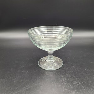 Puede incluir: Un cuenco de pedestal de vidrio transparente con un diseño acanalado. El cuenco está sobre un fondo negro.