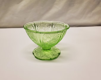 May include: A green glass dessert dish with a pedestal base. The bowl has a scalloped rim and is decorated with a floral pattern. The dish is approximately 4 inches tall and 4 inches wide.