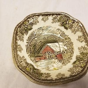 May include: A white ceramic bowl with a green and brown floral pattern. The bowl features a scene of a covered bridge in a snowy winter landscape.
