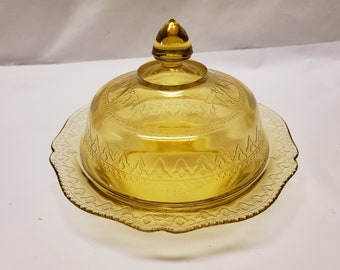 May include: Amber-colored glass butter dish with a domed lid and a scalloped base. The lid features a decorative pattern around the rim and a small knob on top. The base has a matching design. A vintage kitchenware item.