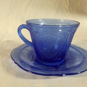 May include: A blue glass teacup and saucer set with an intricate floral pattern. The set is made of clear glass with a blue tint.