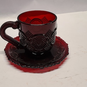 May include: A red glass teacup and saucer set with a decorative pattern. The cup and saucer are both made of ruby red glass and have a raised design.