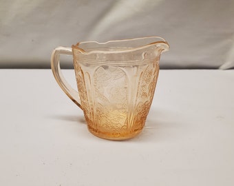 May include: A vintage, peach-colored glass creamer with a handle and a spout. The creamer features a floral pattern etched into the glass. The creamer is likely from the mid-20th century and is a decorative piece for serving cream or other liquids.