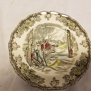 May include: A decorative ceramic bowl with a scalloped edge and a winter scene. The bowl features a central image of a red barn in a snowy landscape, surrounded by a border of green and brown leaves.
