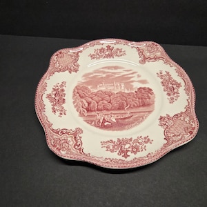 May include: A decorative plate with a scalloped edge, featuring a red and white design. The plate showcases a central landscape scene with a building and trees, framed by floral patterns. The plate is approximately 25 cm in diameter.