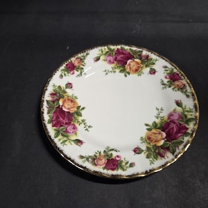 Puede incluir: Un plato de cerámica blanca con borde dorado, adornado con un motivo floral de rosas burdeos y amarillas y hojas verdes. El plato redondo tiene un borde festoneado, ideal para servir postres o aperitivos.