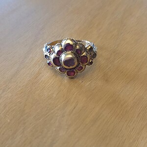 May include: A gold-toned ring with a floral design. The ring features a central cluster of large, round, red gemstones surrounded by smaller red and blue gemstones. The ring is set on a light-colored wooden surface.