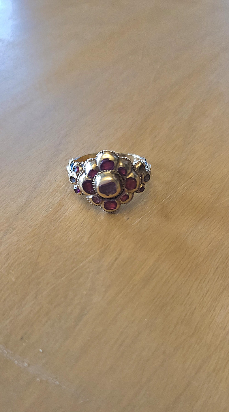 May include: A gold-toned ring with a floral design. The ring features a central cluster of large, round, red gemstones surrounded by smaller red and blue gemstones. The ring is set on a light-colored wooden surface.