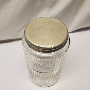 May include: Clear glass jar with a ribbed design and a metal lid. The jar has a cylindrical shape and a wide opening. The lid is round and appears to be made of a silver-colored metal. The jar is empty.