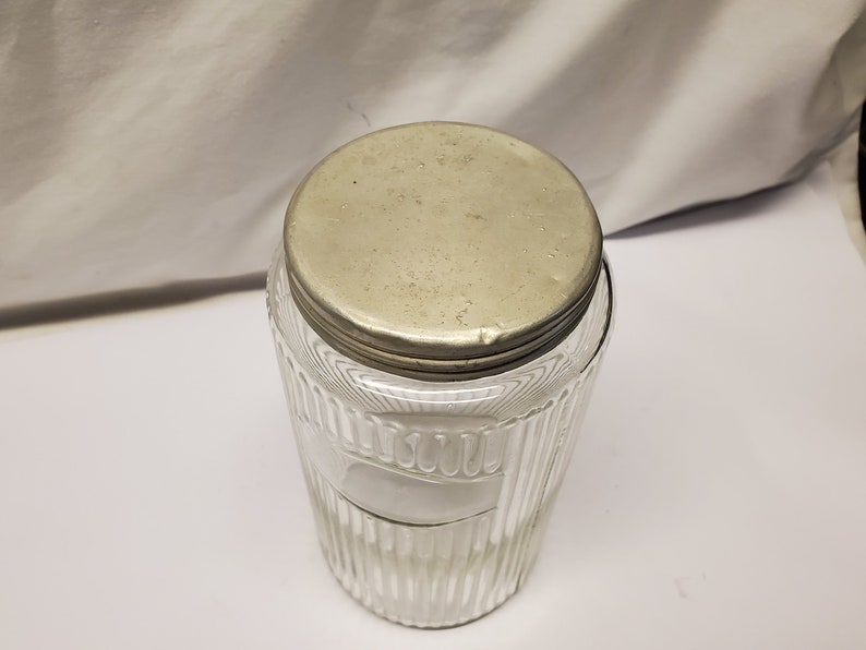 May include: Clear glass jar with a ribbed design and a metal lid. The jar has a cylindrical shape and a wide opening. The lid is round and appears to be made of a silver-colored metal. The jar is empty.
