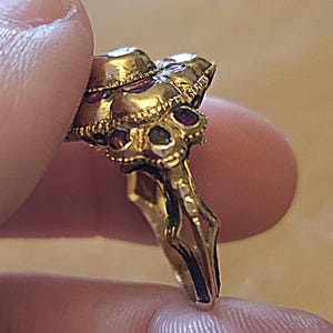 May include: A gold-toned ring with a cluster of gemstones. The ring features a detailed design with multiple small, red gemstones and larger, clear stones. The ring is held between fingers, showcasing its intricate craftsmanship and vintage style.