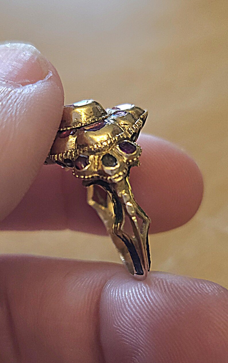 May include: A gold-toned ring with a cluster of gemstones. The ring features a detailed design with multiple small, red gemstones and larger, clear stones. The ring is held between fingers, showcasing its intricate craftsmanship and vintage style.