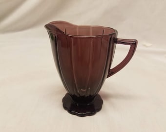 May include: A small, purple glass creamer with a fluted design and a handle. The creamer has a scalloped base.