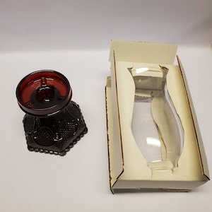 May include: A deep red glass pedestal bowl featuring a textured hobnail pattern on its exterior and a smooth circular rim. Beside the bowl is a rectangular white cardboard packaging insert with a die-cut opening designed to hold the glass object.