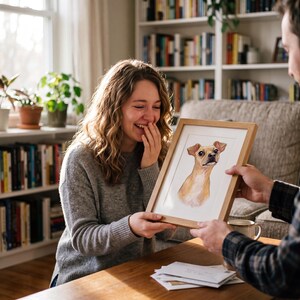 May include: A framed watercolor portrait of a tan and brown dog is being presented to a woman. The portrait is in a light wood frame. The woman is smiling and laughing. The background includes a bookshelf and a window.
