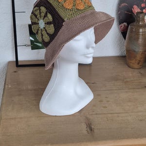 Op de afbeelding: Een bruine gehaakte emmerhoed met een brede rand en een kleurrijk bloemenontwerp. De hoed is versierd met drie gehaakte bloemen in tinten groen, bruin, oranje en wit.