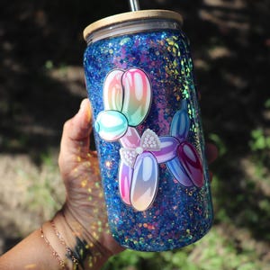 May include: A blue glitter tumbler with a cartoon balloon dog design. The tumbler has a wooden lid and a straw. The balloon dog is in shades of pink, blue, and purple with a white bow. The tumbler is cylindrical.