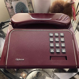May include: A burgundy-colored vintage telephone with a rectangular base and a curved handset. The phone has a keypad with numbered buttons and the word "Diplomat" printed on the front. The phone is sitting on a glass table.