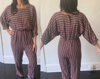 May include: A striped jumpsuit with a brown and purple color scheme. The jumpsuit has long sleeves and a round neckline. The model is wearing black sneakers.