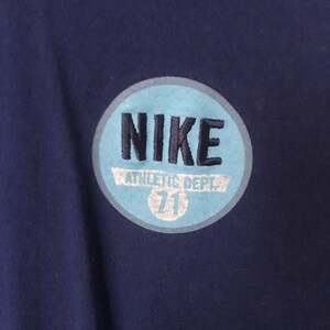 以下が含まれることがあります： 紺色のTシャツに、薄い青色の円形のワッペンが付いています。ワッペンには黒い文字で「NIKE」と、白い文字で「ATHLETIC DEPT. 71」と書かれています。