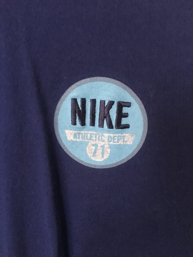 以下が含まれることがあります： 紺色のTシャツに、薄い青色の円形のワッペンが付いています。ワッペンには黒い文字で「NIKE」と、白い文字で「ATHLETIC DEPT. 71」と書かれています。