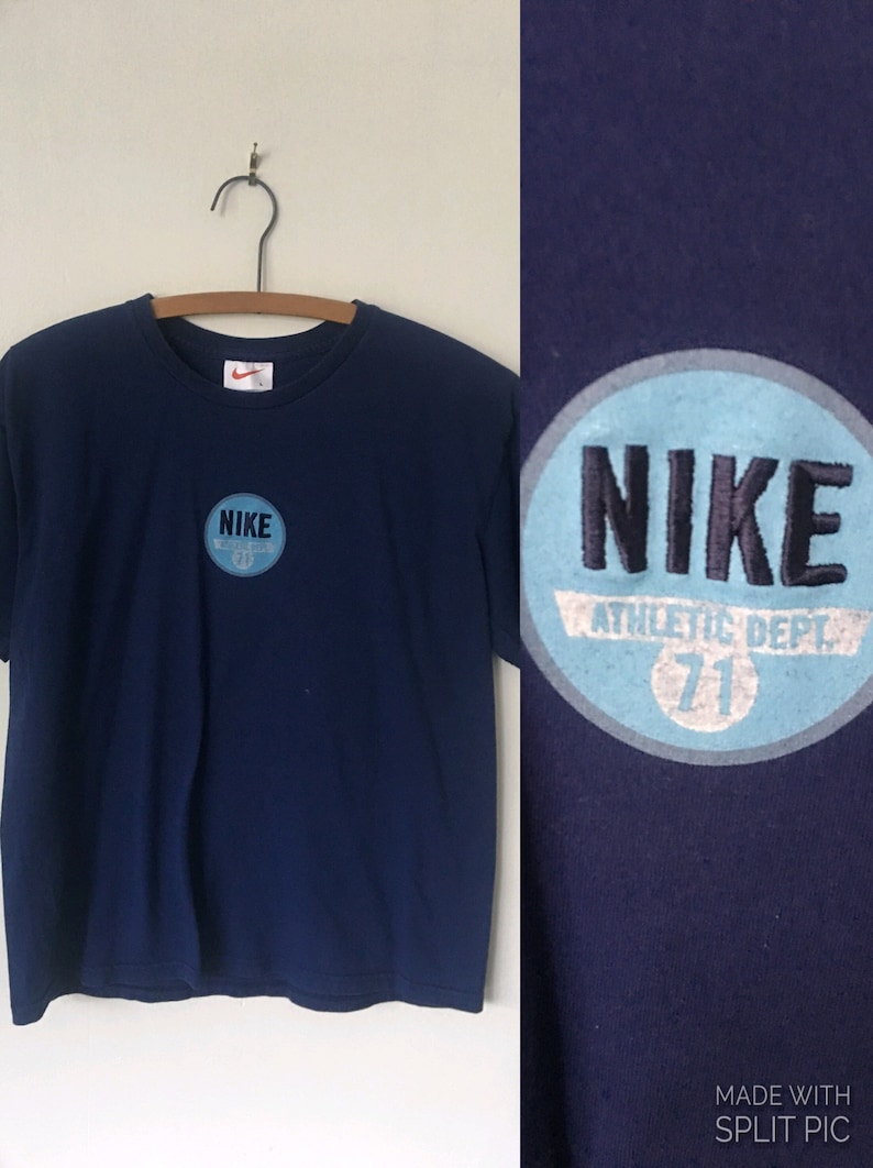 以下が含まれることがあります： 紺色のTシャツに、青と白の円形のロゴがあり、「NIKE ATHLETIC DEPT. 71」と書いてあります。