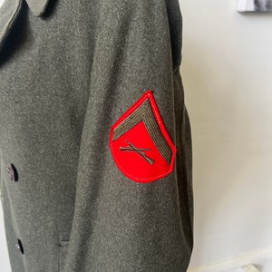 Może przedstawiać: Zielony wełniany płaszcz wojskowy z czerwoną naszywką na rękawie. Naszywka przedstawia skrzyżowane karabiny i szaron.