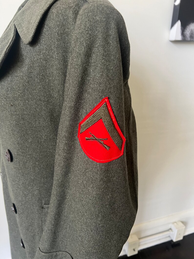 Może przedstawiać: Zielony wełniany płaszcz wojskowy z czerwoną naszywką na rękawie. Naszywka przedstawia skrzyżowane karabiny i szaron.