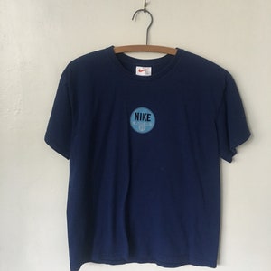 以下が含まれることがあります： 紺色のTシャツに、薄い青色の円形のワッペンが付いています。ワッペンには白い文字で「NIKE」と書いてあり、小さな白いスウッシュロゴも付いています。