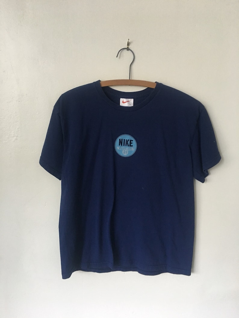 以下が含まれることがあります： 紺色のTシャツに、薄い青色の円形のワッペンが付いています。ワッペンには白い文字で「NIKE」と書いてあり、小さな白いスウッシュロゴも付いています。