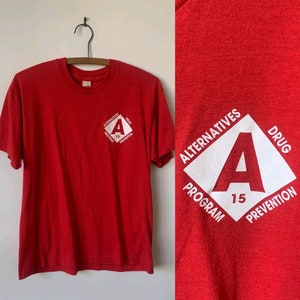 May include: A red t-shirt with a white diamond-shaped graphic on the front. The graphic includes the letter "A", the number "15", and the words "Alternatives", "Drug", "Program", and "Prevention". The shirt is hanging on a wooden hanger.