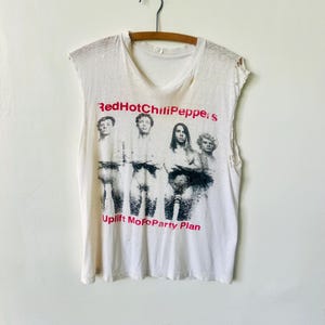 May include: White sleeveless t-shirt featuring a black and white image of the band Red Hot Chili Peppers. The band's name is printed in red and the text "Uplift Mofo Party Plan" is printed below the image.