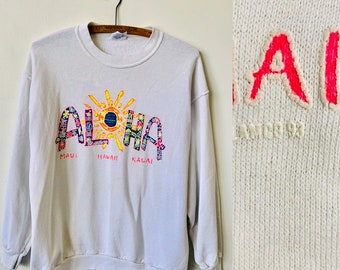 Op de afbeelding: Witte trui met een kleurrijke "Aloha" afbeelding met een zon en de woorden "Maui", "Hawaii" en "Kalai".